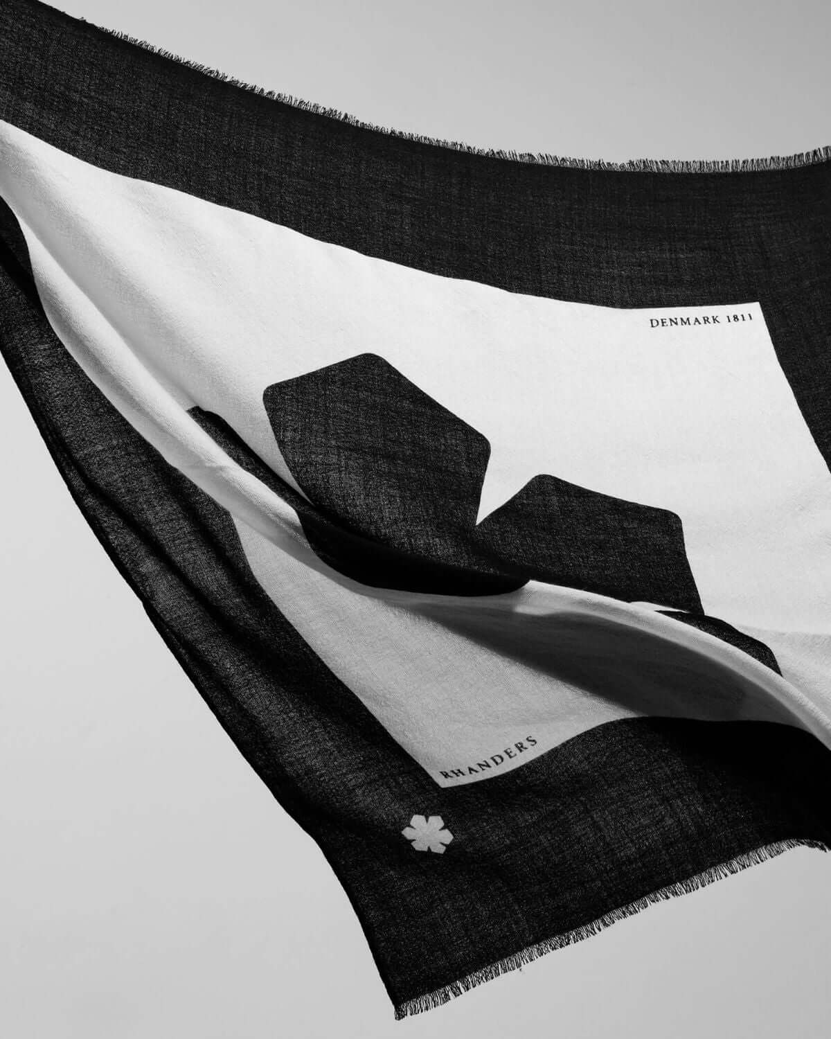 The Adora Scarf in the colour black / champagne adorned with a grand, centered Kalmus flower and a coloured frame featuring our Kalmus flower, RHANDERS, and DENMARK 1811.